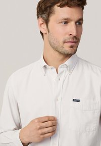 Camisa blanca de algodón con cuello abotonado, un bolsillo en el pecho y una etiqueta de marca en color marino. Textura suave y diseño clásico.