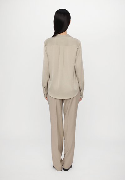 Beige long-sleeve blouse with a rounded hem and central pleat, paired with high-waisted beige trousers, featuring a tailored fit and smooth texture.