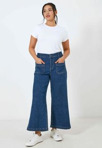 Weite, dunkelblaue Jeans mit zwei Fronttaschen, kombiniert mit einem weißen T-Shirt mit kurzen Ärmeln und weißen Sneakers, vor einem neutralen Hintergrund.