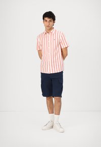 Kortermet skjorte med knappestramming i røde og hvite vertikale striper, kombinert med marineblå cargo-shortser og hvite joggesko, stående mot en ensfarget bakgrunn.