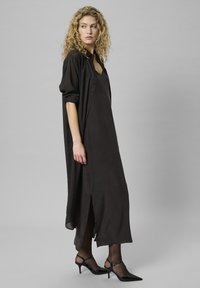 Femeie blondă îmbrăcată într-o rochie lungă neagră fără mâneci, cu slit lateral și cămașă neagră transparentă cu nasturi, purtând ciorapi negri transparenți și pantofi cu toc ridicat.