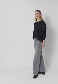 Pull noir en maille à col rond associé à un jean évasé gris clair. Le mannequin porte des bottines noires. Arrière-plan simple et neutre.