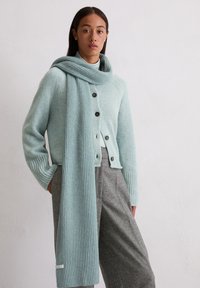 Cardigan tricoté bleu clair avec des manches côtelées, devant à boutons et écharpe assortie. Pantalon gris avec des plis et une texture lisse.