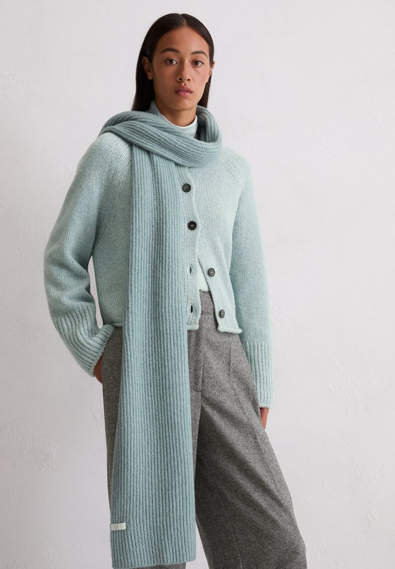 Cardigan tricoté bleu clair avec des manches côtelées, devant à boutons et écharpe assortie. Pantalon gris avec des plis et une texture lisse.