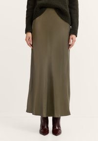 Olivenfarbene Satin-Maxiröcke mit glatter Textur, die eine gerade Silhouette und einen seitlichen Schlitz aufweisen, kombiniert mit dunkelbraunen spitzen Stiefeln.