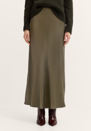 Olivenfarbene Satin-Maxiröcke mit glatter Textur, die eine gerade Silhouette und einen seitlichen Schlitz aufweisen, kombiniert mit dunkelbraunen spitzen Stiefeln.