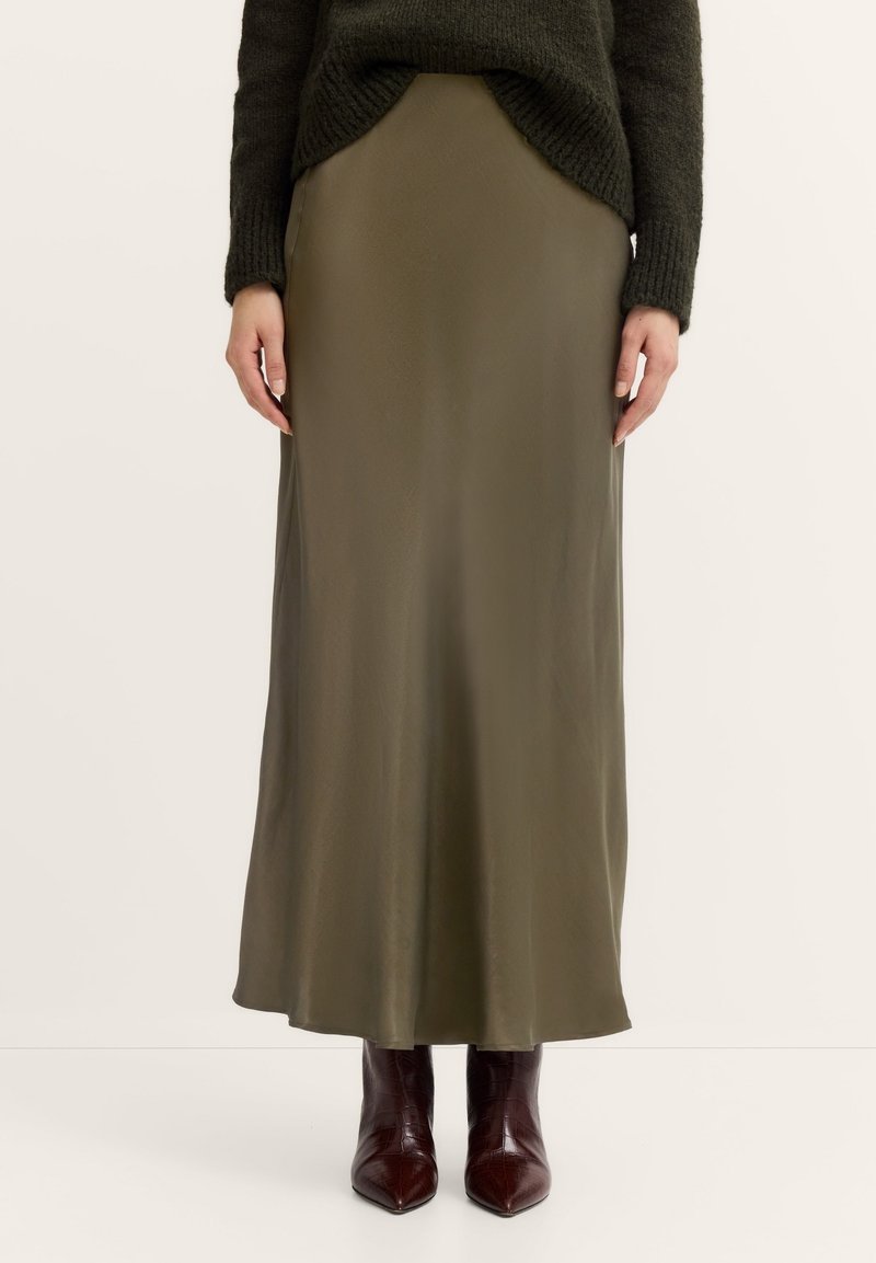 Olivenfarbene Satin-Maxiröcke mit glatter Textur, die eine gerade Silhouette und einen seitlichen Schlitz aufweisen, kombiniert mit dunkelbraunen spitzen Stiefeln.