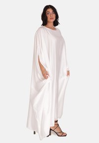 Witte satijnen kaftan jurk met een losse pasvorm, ronde halslijn en zijsplitten. Gepaard met zwarte sandalen met banden voor contrast.