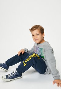 Sudadera gris con el texto colorido "MARVEL", combinada con pantalones de chándal azul marino que tienen texto amarillo. Zapatillas azules completan el atuendo.