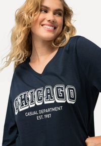 Marineblaues Langarm-T-Shirt mit V-Ausschnitt, das auf der Vorderseite in fetten weißen Buchstaben BBCHICAGO CASUAL DEPARTMENT EST. 1987AB zeigt. Glatte Textur.