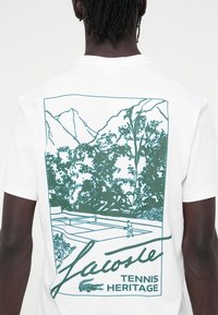 Camiseta de algodón blanca con un gráfico verde en la parte posterior que representa montañas, árboles, una pista de tenis y el texto "Lacoste Tennis Heritage".