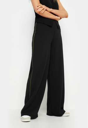 Pantaloni neri a gamba larga con una texture liscia e una striscia laterale verde a contrasto, abbinati a sneakers bianche per un look casual.