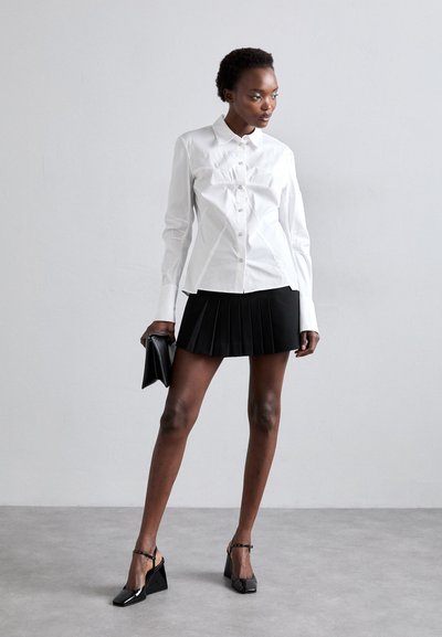 White button-up shirt with vertical seams, paired with a black pleated mini skirt. Model wears black patent slingback shoes and holds a clutch.
