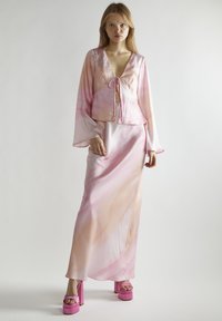 Rosa Satin-Outfit mit einem kurzen Oberteil mit Krawattenfront und langen Ärmeln, kombiniert mit einem knöchellangen Rock. Hohe rosa Plateau-Sandaletten.