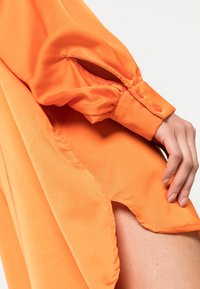 Blusa naranja de tejido suave, con mangas largas y cierre de botón, y aberturas laterales para mayor movimiento y transpirabilidad.