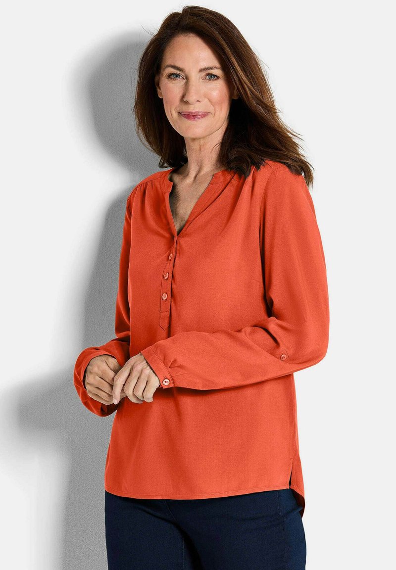 Blusa arancione brillante con scollatura a V, patta con bottoni frontale, maniche lunghe e spacchi laterali. Texture liscia e tessuto morbido.