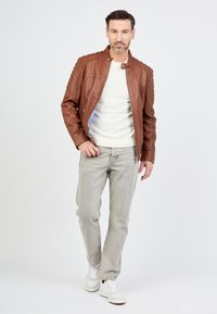 Veste en cuir marron avec des détails matelassés aux épaules, portée sur un pull gris clair et assortie à un jean gris et des baskets blanches.