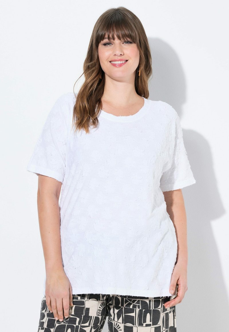Top bianco a maniche corte con texture floreale e scollatura tonda. Il tessuto sembra morbido, abbinato a pantaloni neri a fantasia con disegni astratti.