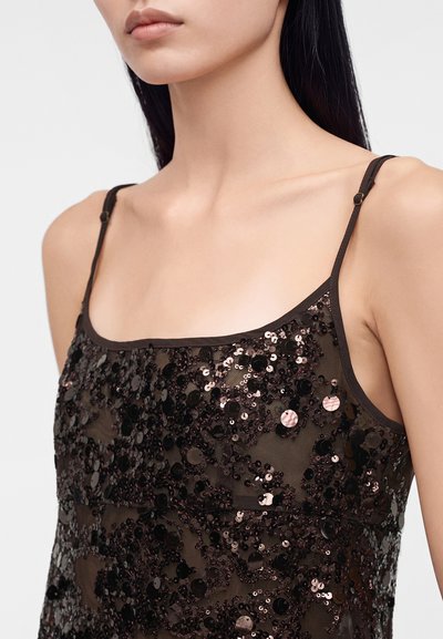 Femme portant un haut noir transparent sans manches orné de sequins noirs et foncés brillants, montrant la zone du cou et des épaules.