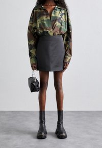 Donna che indossa una camicia a maniche lunghe mimetica, una mini gonna nera, stivali chunky neri e che porta una piccola borsa nera con manico a catena.