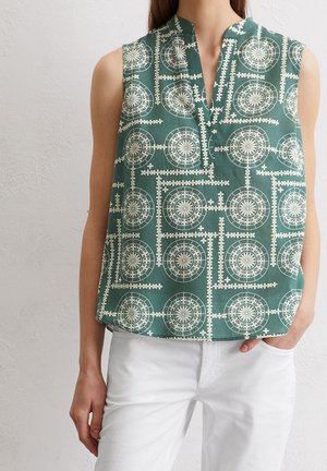 Ärmelloses grünes Top mit weißen kreisförmigen und floralen geometrischen Mustern, getragen mit weißen Hosen, Hand in der Tasche, vor einer hellen Wand stehend.