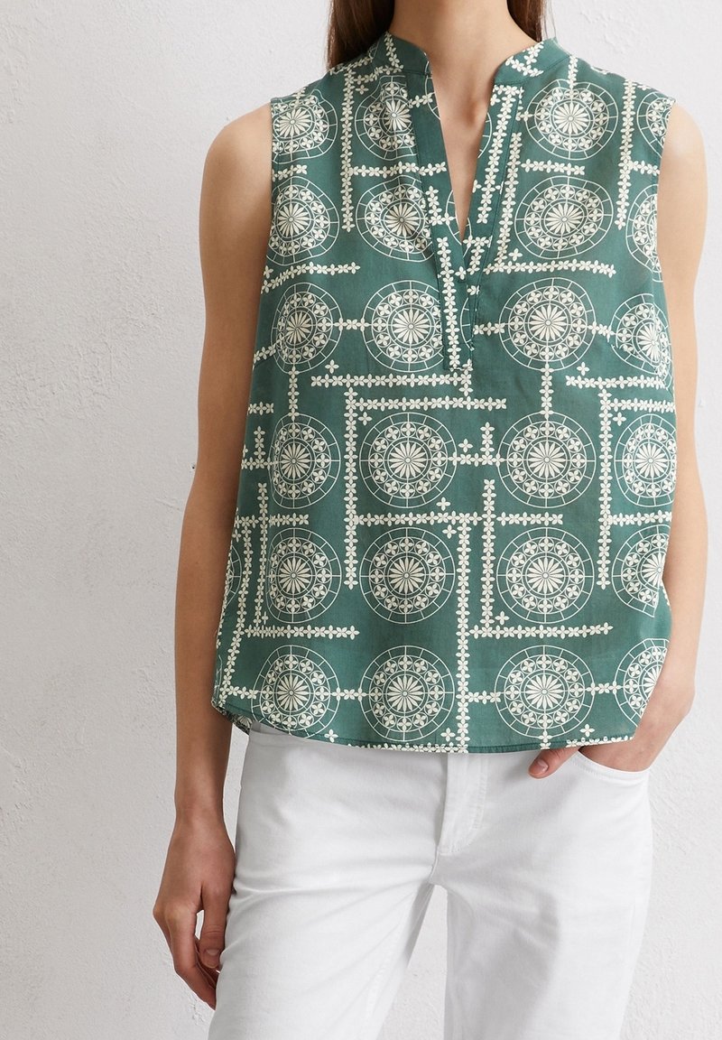 Haut vert sans manches avec des motifs géométriques circulaires et floraux blancs, porté avec un pantalon blanc, main dans la poche, debout contre un mur clair.