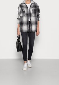 Chaqueta de franela a cuadros en blanco y negro con dos bolsillos en el pecho, llevada sobre una camiseta blanca, combinada con jeans Skinny oscuros y zapatillas blancas.