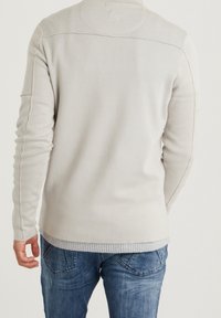 Hellgrauer Pullover mit geripptem Saum und verlängerten Ärmeln, der sichtbare Nahtakzente an den Schultern und Armen aufweist. Kombiniert mit blauen Jeans.