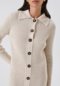 Beige ribgebreide cardigan met een brede kraag, vijf bruine knopen en lange mouwen. Het materiaal heeft een zachte, gestructureerde uitstraling.