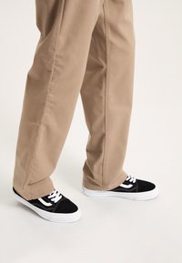 Sneaker basse bianco e nero con lacci, abbinate a pantaloni dritti color beige. Le sneaker presentano una texture in suede e tela.