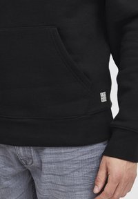 Black sweatshirt featuring a front pocket, ribbed cuffs, and a small logo tag. The fabric appears soft with a smooth texture.