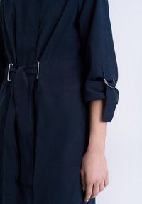 Robe bleu marine avec taille ceinturée, présentant une texture lisse, des manches retroussées et des accents de quincaillerie argentée. Tissu doux et coutures minimales.