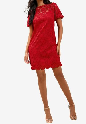 Femme portant une courte robe rouge en dentelle avec des manches bouffantes et un ourlet festonné, associée à des sandales à talons hauts beiges, sur un fond blanc.