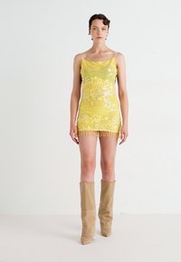 Frau in einem gelben, floral bestickten Minikleid mit Fransensaum und beigen kniehohen Stiefeln, die vor einem schlichten weißen Hintergrund steht.