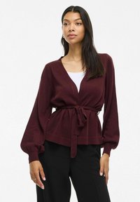 Cardigan bordeaux a V, con maniche lunghe a palloncino e cintura in vita. Tessuto a maglia testurizzato. Abbinato a una maglietta bianca e pantaloni neri.