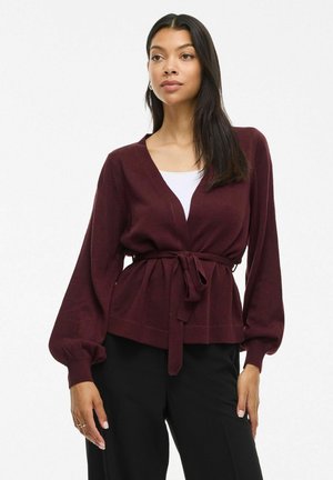 Bordeauxfarbener V-Ausschnitt-Cardigan mit langen, ballonförmigen Ärmeln und einem taillierten Bindegürtel. Aus strukturiertem Strickstoff. Kombiniert mit einem weißen Oberteil und schwarzen Hosen.