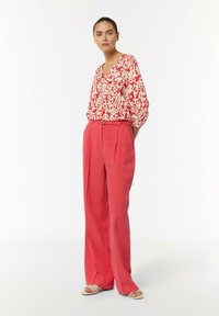 Czerwona i biała bluzka w wzory z dekoltem w serek, zestawiona z wysokimi koralowymi spodniami o szerokich nogawkach. Całość dopełniają płaskie sandały.