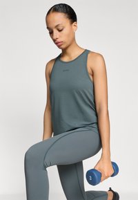 Zaļš sporta topiņš ar apaļu kakla izgriezumu, pārī ar atbilstošām legingām. Subjekts tur zilu hanteli ar atzīmi "3".