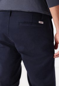 Pantaloni in misto cotone blu navy con taglio dritto, tasca posteriore, etichetta con logo e texture liscia. Presentano una doppia cucitura e passanti per la cintura.