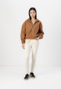 Veste zippée marron à coupe oversize, avec deux poches avant et un ourlet côtelé. Associée à un pantalon skinny couleur crème et des baskets noires.
