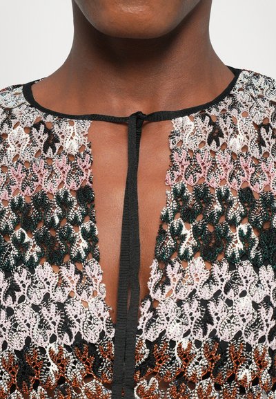 Close-up of a person wearing a multicolored crocheted top with black trim and a black tie at the neckline.
