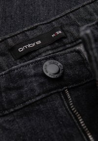 Pantalon en denim foncé avec une étiquette "ombre". Il comporte un bouton noir et des coutures contrastées le long du bord. Tissu texturé avec une finition lisse.