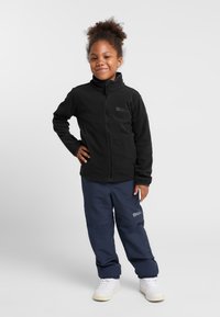Veste en polaire noire avec col haut, fermeture éclair et logo ; associée à un pantalon bleu marine foncé et des baskets blanches. L'enfant se tient debout avec confiance.
