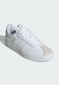 White sneaker with a textured leather upper, suede toe cap, and three stripes in gold. Features a padded collar and a rubber sole.