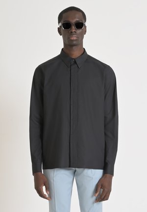 Camicia nera a maniche lunghe con colletto classico, chiusura con bottoni frontale e texture liscia, abbinata a pantaloni azzurri chiari e occhiali da sole.