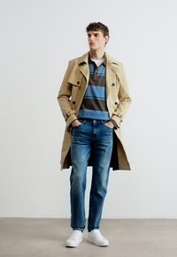 Νεαρός άνδρας με μπεζ αντιανεμικό παλτό, μπλε ριγέ πόλο πουκάμισο, τζιν και λευκά αθλητικά παπούτσια, στέκεται με τα χέρια στις τσέπες μπροστά σε λευκό τοίχο.