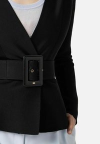 Blazer nero con cintura indossato sopra top bianco e pantaloni bianchi, con la mano destra appoggiata in tasca su sfondo neutro.