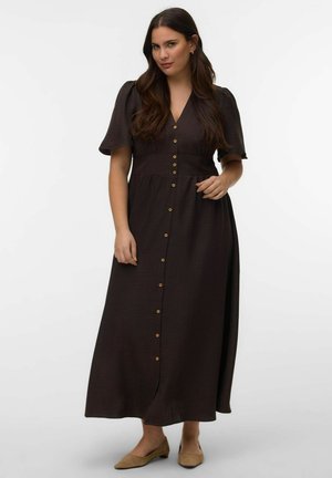 Ženska z dolgimi temnimi lasmi, oblečena v črno midi obleko z gumbi spredaj in napihnjenimi rokavi ter bež ravnimi čevlji, stoji na belem ozadju.