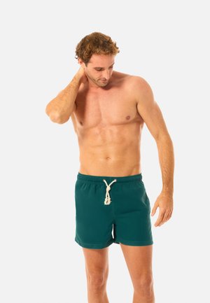 Paita pois päältä oleva mies, jolla on päällään petroolinsiniset uimashortsit, joissa on valkoinen nyöri, katsoo alas ja koskettaa niskaansa seisten yksinkertaisen vaalean taustan edessä.