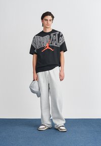 Un tânăr îmbrăcat cu un tricou negru Jordan, pantaloni de trening gri, pantofi sport multicolori, ținând o șapcă gri, stând pe un covor bleu.